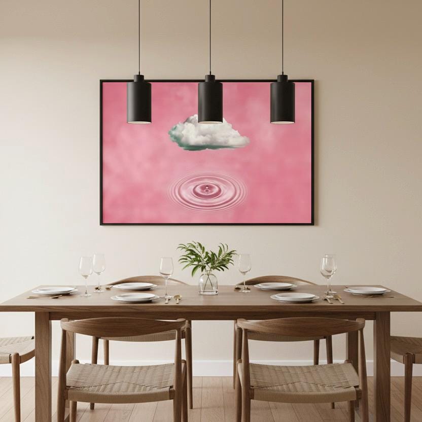 a dining room with a wooden table set for four, featuring white plates and wine glasses. Above the table, there is a framed artwork depicting a pink background with a cloud and a ripple effect. The artwork is hung on a white wall, and three black pendant lights are suspended above the table.