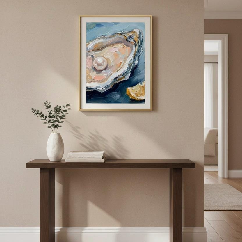 a framed painting of an oyster shell with a pearl, hanging on a wall above a wooden console table with a vase of greenery and a stack of books.